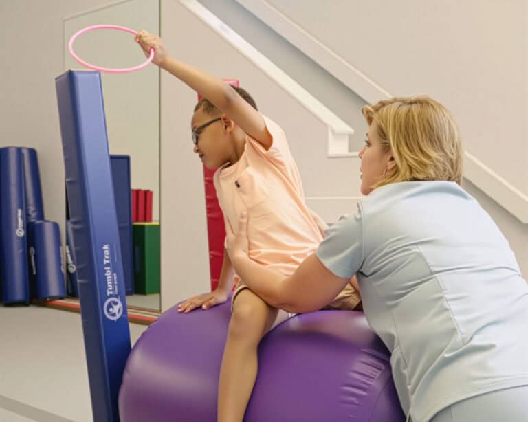 A photo of a young client doing a therapeutic exercise on a Air Barrel with rings and a Fun Stick, guided by an occupational therapist.