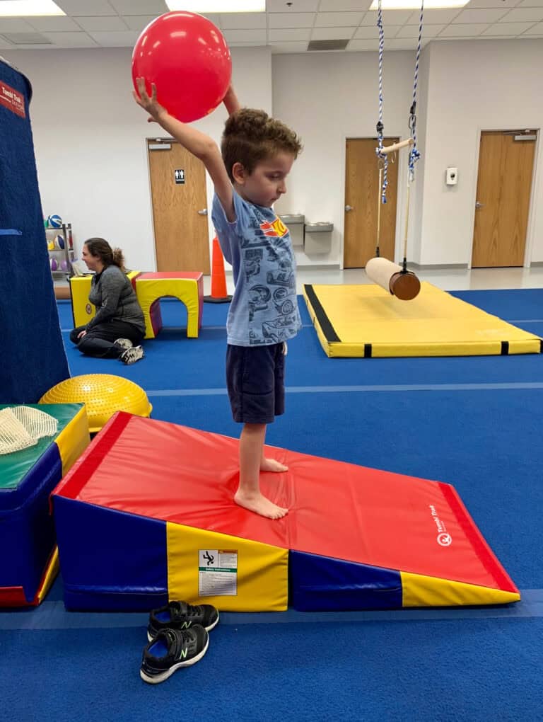 A photo of a boy in an occupational therapy clinic standing on a Folding Incline Mat and holding a ball.