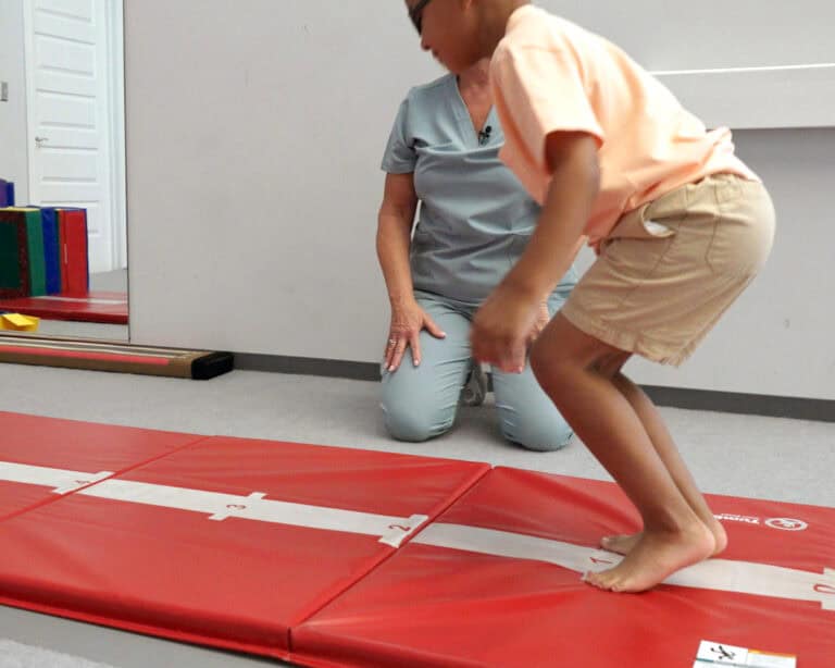 A photo of a boy jumping on a Hopscotch Mat while his occupational therapist watches.