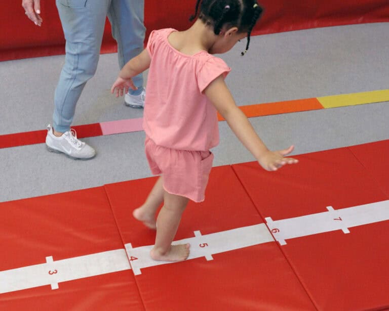 A photo of a girl walking the center line on a Hopscotch Mat while her occupational therapist watches.