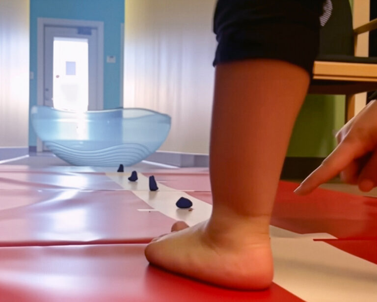 A photo showing small objects placed on the white line of the Hopscotch Mat. In the foreground, we see the feet of an occupational therapy client as they prepare to begin the exercise.
