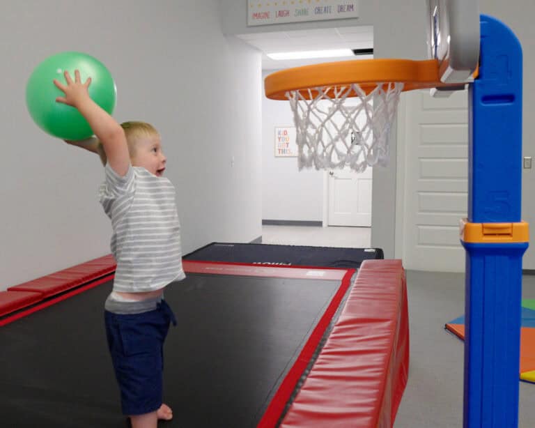 A photo of a boy standing on a Tumbl Trak while about to toss a therapy ball into a toy basketball hoop.