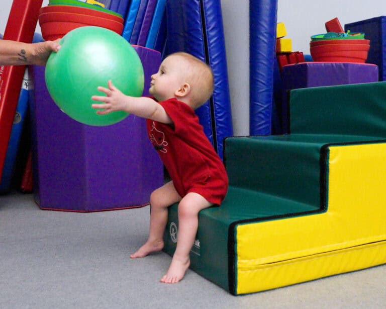 A photo of a baby sitting on Tumbl Trak Steps being handed a balloon by an occupational therapist.