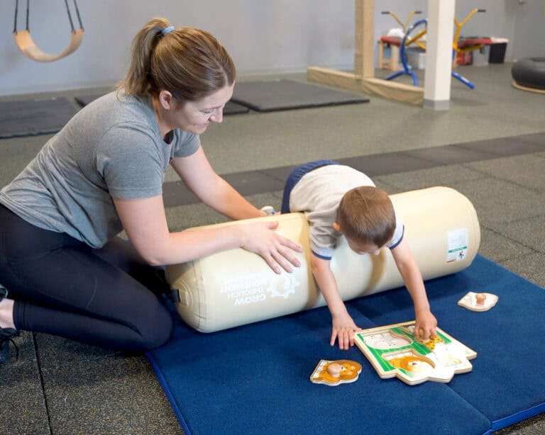A photo of an occupational therapist working with a boy on the Air Bolster Mini. He is resting across it reaching for manipulatives.
