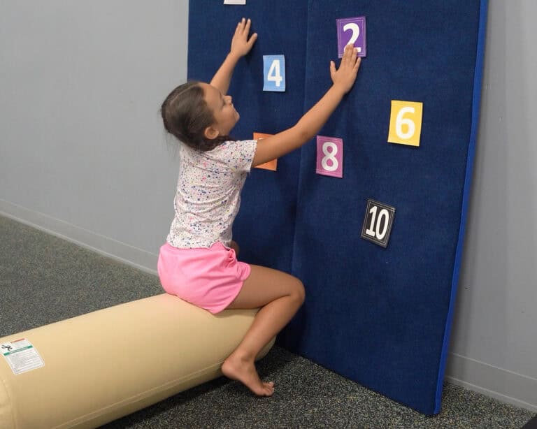 A photo of a girl sitting on an Air Bolster Mini and placing Sticky Manipulatives onto a velcro wall.