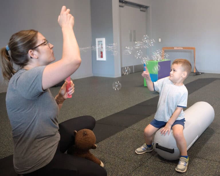 A photo of a boy sitting on an Air Bolster Mini as his occupational therapist blows bubbles.